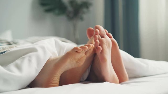 Human bare feet male and female are touching each other in bed under white blanket on sheets in bedroom. Happiness, intimate relationship and people concept.