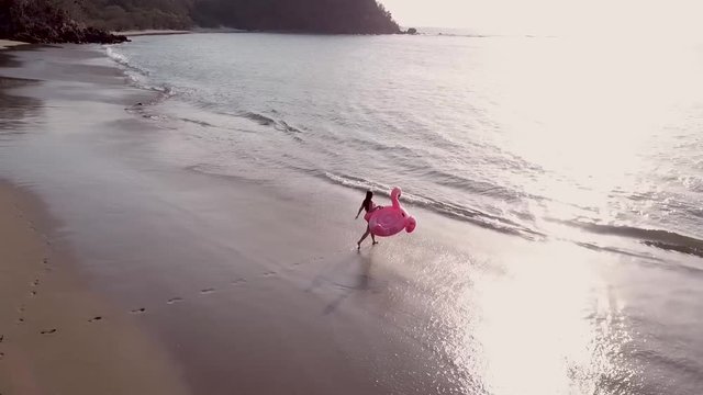 Young woman walking towards the sea holding a floating flamingo at sunset.