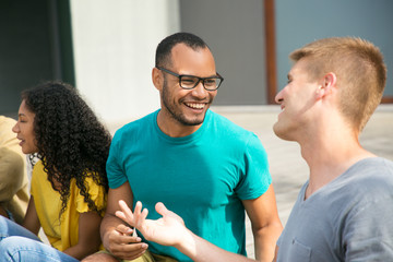Happy male friends chatting and having fun outside. Caucasian and mix raced guys sitting outdoors, talking, laughing at joke. Friends having fun concept