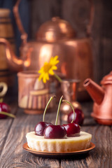 Small portion cheesecake with fresh sweet cherries against of cooper dish and scattered sweet cherries over on old wooden background. Rustic style.