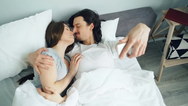 Husband and wife happy young people are taking selfie with smartphone camera kissing hugging lying in bed together. Home, youth and love concept.