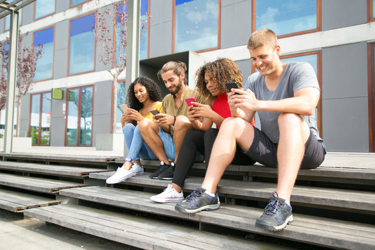 Happy multiethnic students sitting together and using their cellphones. Interracial group of people sitting outside, holding smartphones, using online app, texting message. Communication concept