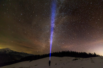 Back view of man with head flashlight standing on snowy valley under beautiful dark blue winter starry sky, bright blue beam on stars and mountains ridge background. Night photography, light painting.