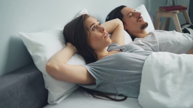 Irritated woman is covering head with pillow when boyfriend is snoring in sleep in bed disturbing her relaxation. Healthcare, problems and family concept.