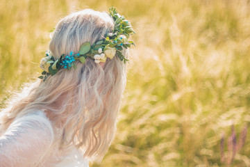 Back of the young woman in a white dress in boho style with a floral wreath in the summer in the field.
