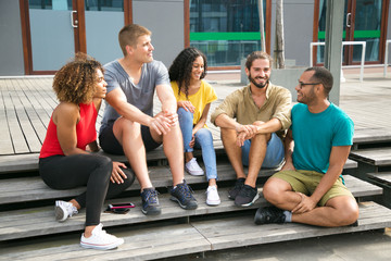 Team of cheerful mix raced friends sharing funny stories. Interracial group of young people sitting on outdoor staircase, talking, listening, laughing. Happy meeting concept