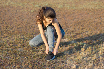 A slim athletic girl sits, tying shoelaces on sneakers in nature.