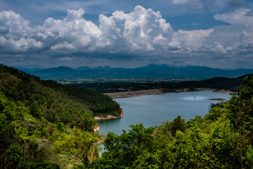 View of the reservoir surrounded by mountains and green forest from the mountain top against cloudy sky with white fluffy cumulus clouds.
