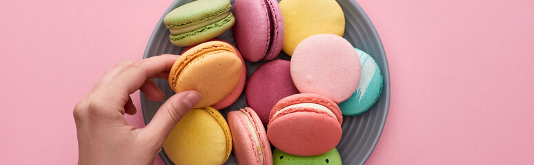 cropped view of female hand near plate with multicolored delicious French macaroons on pink background, panoramic shot