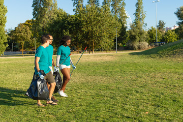 Mix raced team of volunteers cleaning city park from garbage. Young woman and men walking on grass, carrying rakes and plastic bags. Trash removal concept