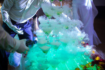 waiters fill martini glasses drawn up by a slide, green cocktail cherries in glasses, steam from dry ice above glasses