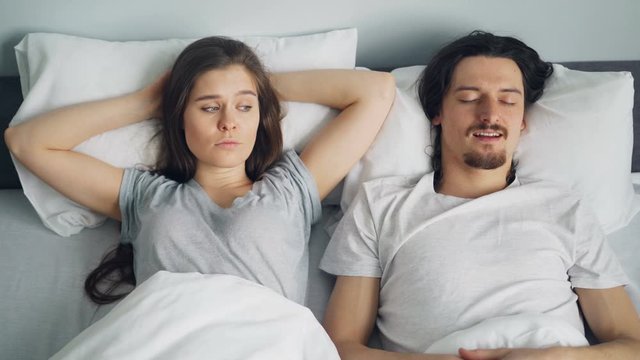 Angry wife attractive girl is covering face with pillow while sleeping husband bearded guy is snoring in bed. Family life, problems and bedtime concept.