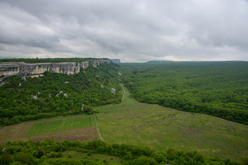 view of the valley of green with trees and grass, on a cloudy day with clouds in the sky, and mountains with flat peaks of white and gray.
