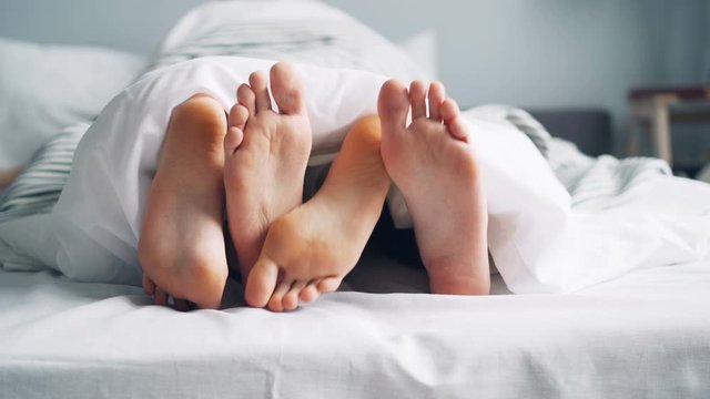 Mans and womans feet are moving under blanket caressing touching in bed on white sheets, people aare enjoying bedtime together. Relationship and body concept.