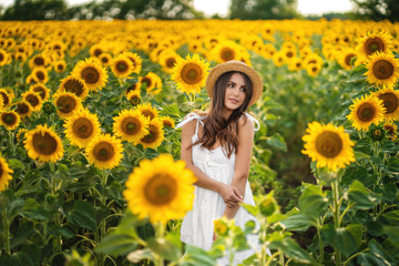 Fototapeta na wymiar Beautiful girl in straw hat and white sundress on a walk by field of sunflower