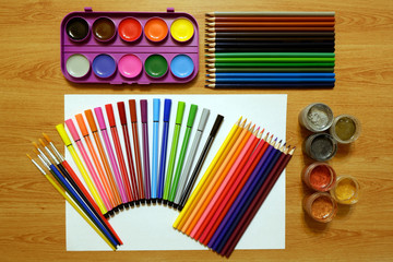 Sets of watercolor paints, acrylic metallic paints and brushes, colored pencils and felt pens, blank sheet of withe paper on a wooden table