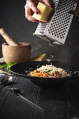 Hands grated Parmesan cheese in spaghetti Bolognese pasta with dried tomatoes and sauce in a black plate on a black wooden background.