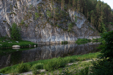 beautiful landscape, the rock is reflected in a calm mountain river, around the forest and green grass. Background for design or photo wallpaper.