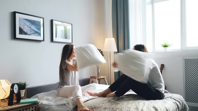 Playful husband and wife are having fun with pillow fight on bed at home laughing and relaxing. Relationship, silly activity and joyful leisure concept.