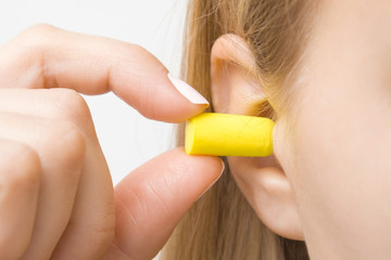 Young woman's fingers putting yellow earplug into her ear on light gray background. Closeup.