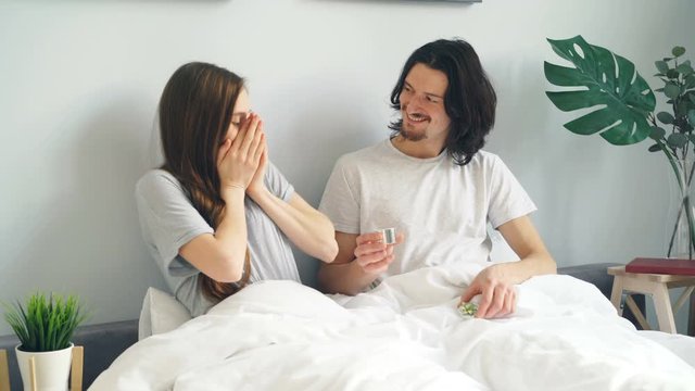 Young man is making marriage proposal to surprised girl putting ring on finger in bed at home laughing kissing hugging. Family, love and relationship concept.
