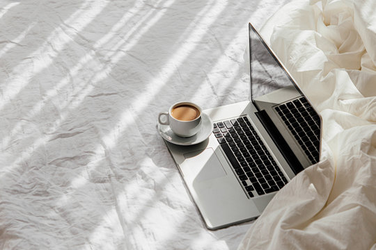 Laptop and cup of coffee on white bed with a  blanket. Work at home concept. Morning light