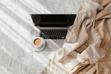 Laptop and cup of coffee on white bed with a beige plaid. Work at home concept. Morning light