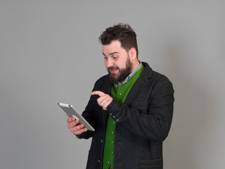 young and cool austrian man with black hair and beard in traditional costume stands in front of a grey background and works on a tablet
