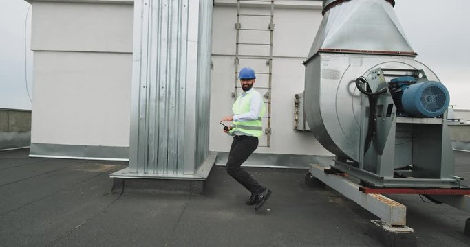 Charismatic and smiling large architect or engineer dancing excited in front of the camera on the top of construction site while holding the speaking ration he wearing a safety helmet