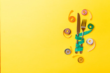 Top view composition of fork and knife with colored balled measuring tapes on yellow background with empty space for your ideas. Overweight and overeating concept