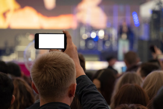 A man is holding a smartphone in his hands and is recording or live broadcasting a street concert among a crowd of fans. View from the back. A blank for embedding any image instead of a white screen.