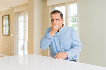 Middle age man sitting at home smelling something stinky and disgusting, intolerable smell, holding breath with fingers on nose. Bad smells concept.