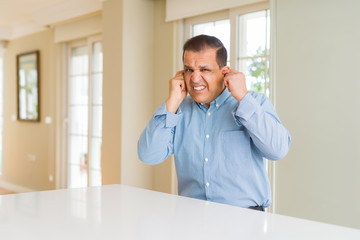 Middle age man sitting at home covering ears with fingers with annoyed expression for the noise of...