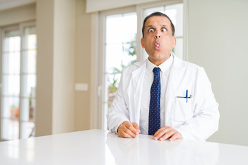 Fototapeta premium Middle age doctor man wearing medical coat at the clinic making fish face with lips, crazy and comical gesture. Funny expression.