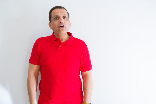 Middle age man wearing red t-shirt over white wall afraid and shocked with surprise expression, fear and excited face.