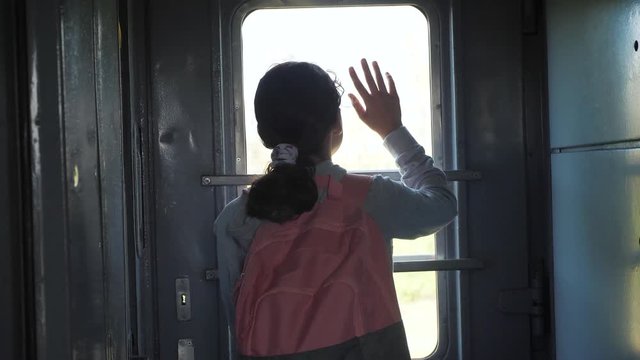little teenage girl is a backpacker traveling by lifestyle train. travel transportation railroad concept. tourist school girl in the train wagon with backpack looking out the window waving his hand