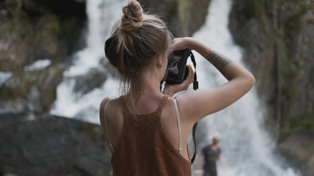 Young paparazzi girl photographs a beautiful large waterfall standing on the stones in the jungle