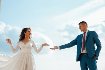 Beautiful woman in a long white dress walks around the castle with her husband in a blue suit on the sky background