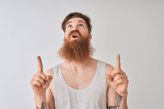 Young redhead irish man wearing t-shirt standing over isolated grey background amazed and surprised looking up and pointing with fingers and raised arms.