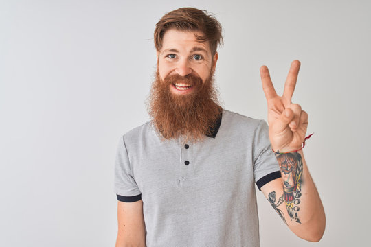Young redhead irish man wearing grey polo standing over isolated white background showing and pointing up with fingers number two while smiling confident and happy.