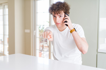 Young man talking on the phone at home pointing with finger to the camera and to you, hand sign, positive and confident gesture from the front