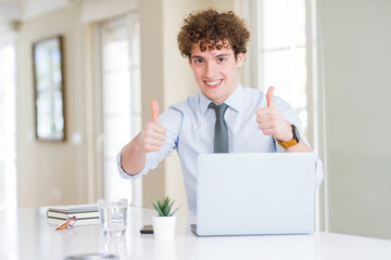 Young business man working with computer laptop at the office approving doing positive gesture with hand, thumbs up smiling and happy for success. Looking at the camera, winner gesture.