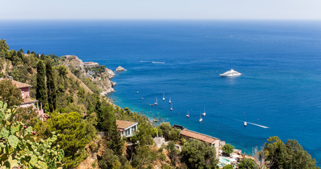 Fototapeta na wymiar Panoramic view from Taormina, in Sicily (Italy). A sunny summer day with beach, blue sky, boats and luxury houses. A cactus can be seen among the green vegetation in the foreground. – Image