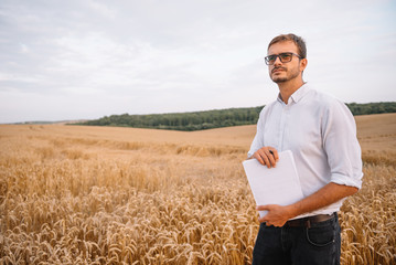 Happy young farmer engineer with notebook standing on wheat field while combine harvester working in background