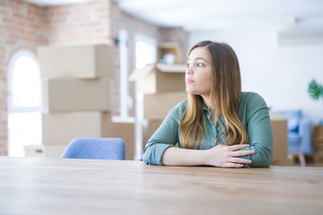 Fototapeta premium Young woman sitting on the table with cardboard boxes behind her moving to new home looking to side, relax profile pose with natural face with confident smile.