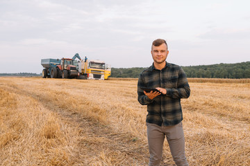 Young agronomist man standing on wheat field checking quality while combine harvester working.