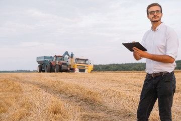 Happy young farmer engineer with notebook standing on wheat field while combine harvester working...