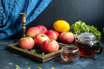 Still life of an old grandmother’s photo frame, apples, a tea pot with a cup of tea and an old metal candlestick. From the back, blue fabric on a dark background. 