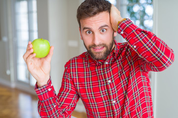 Handsome man eating fresh healthy green apple stressed with hand on head, shocked with shame and surprise face, angry and frustrated. Fear and upset for mistake.
