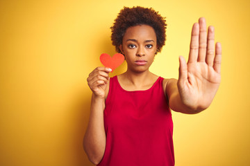 African american woman holding romantic paper hearts over yellow isolated background with open hand doing stop sign with serious and confident expression, defense gesture